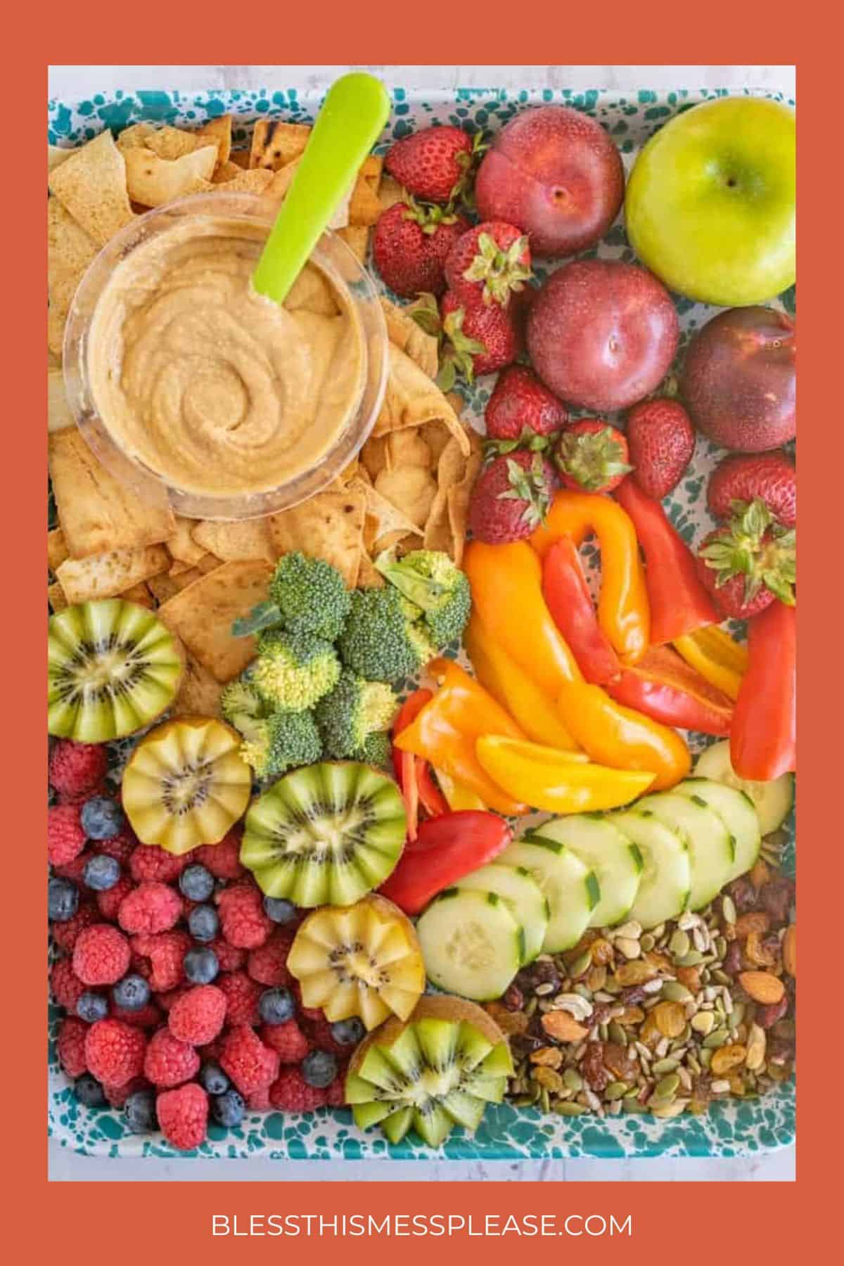 A colorful platter inspired by healthy snack recipes, featuring chips and dip, apple, strawberries, kiwi, raspberries, broccoli, bell pepper slices, cucumber, cherry tomatoes, mixed nuts, and plums on a patterned tray.