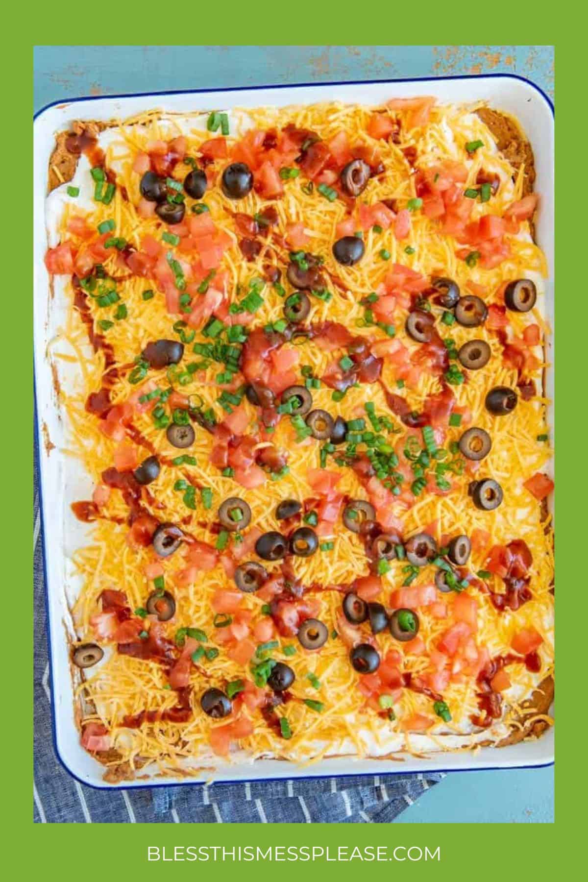 A casserole dish filled with a layered dip topped with shredded cheese, diced tomatoes, sliced black olives, green onions, and bacon bits—an irresistible choice for super bowl appetizers. Surrounded by a green border with at the bottom.