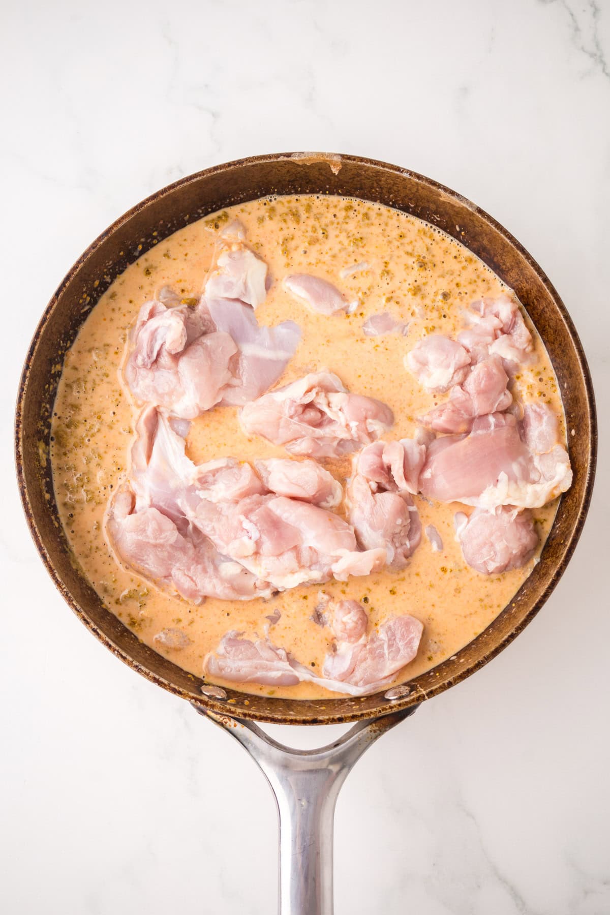 A frying pan filled with raw chicken pieces marinating in a creamy, seasoned sauce sits on a white marble surface.