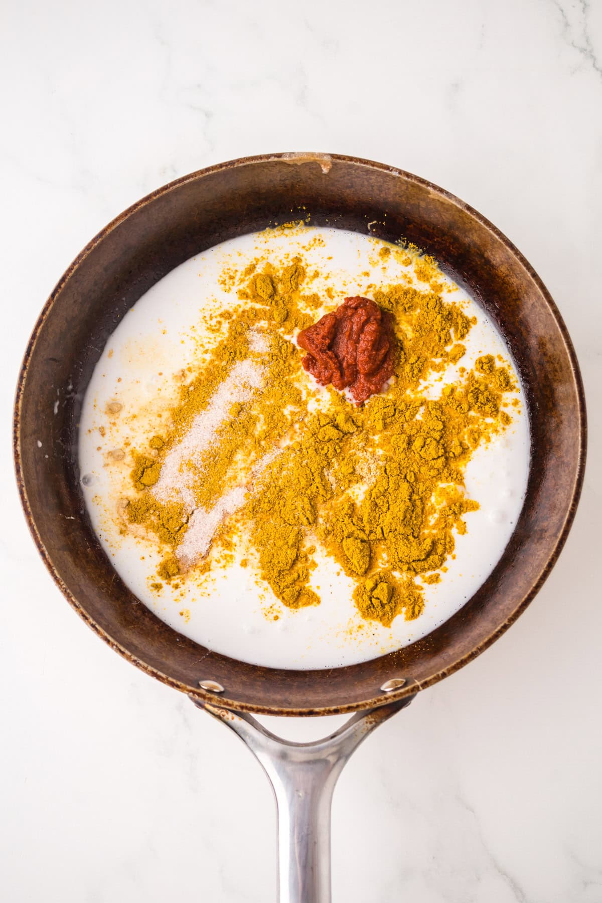 A pan with coconut milk, yellow curry powder, salt, and a dollop of red curry paste sits on a white marble surface, ingredients not yet mixed.