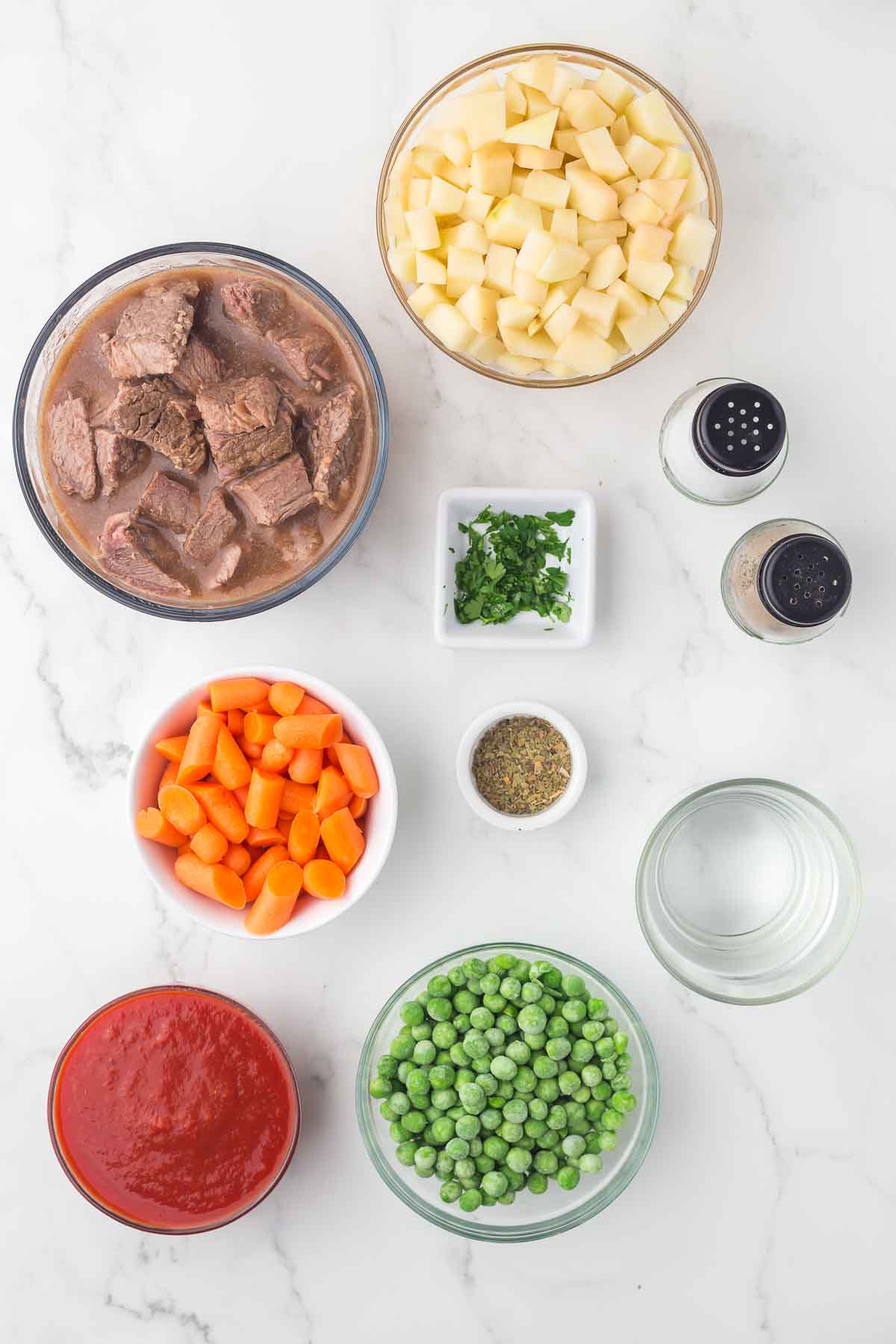 Ingredients for a beef stew recipe arranged on a white surface: cubed beef in broth, diced potatoes, baby carrots, green peas, tomato sauce, water, chopped parsley, dried herbs, salt, and pepper shakers.