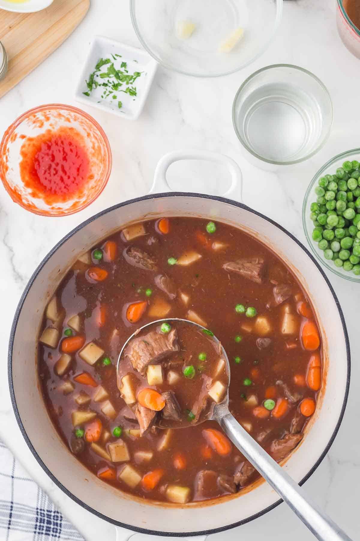 A pot of beef stew with chunks of beef, carrots, potatoes, and green peas. A ladle is dipping into the stew. Ingredients like peas, water, and chopped herbs are in bowls nearby on a white countertop.