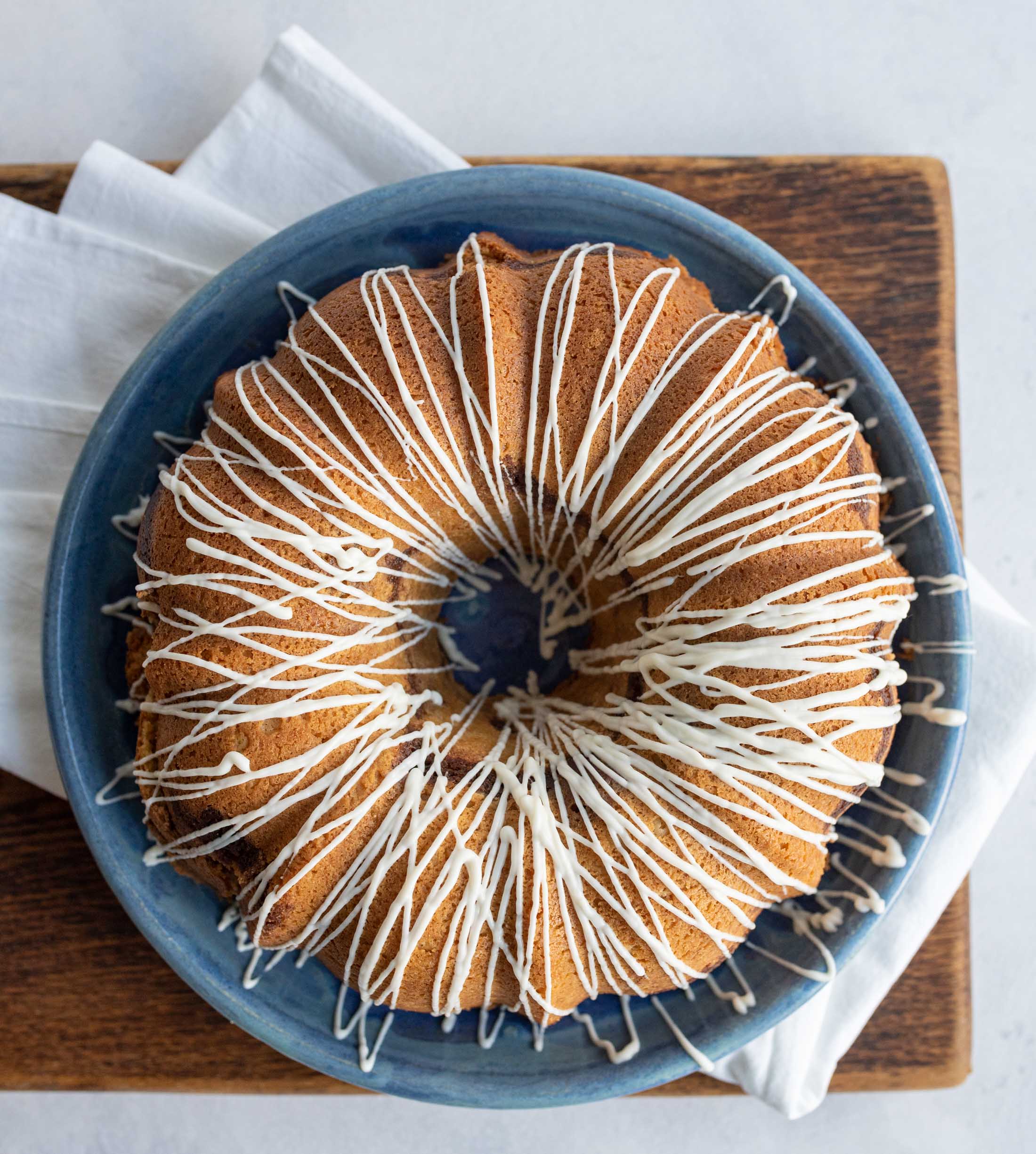 A round bundt cake with white icing drizzled in a zigzag pattern sits on a blue plate, placed on a wooden board with a white napkin underneath.