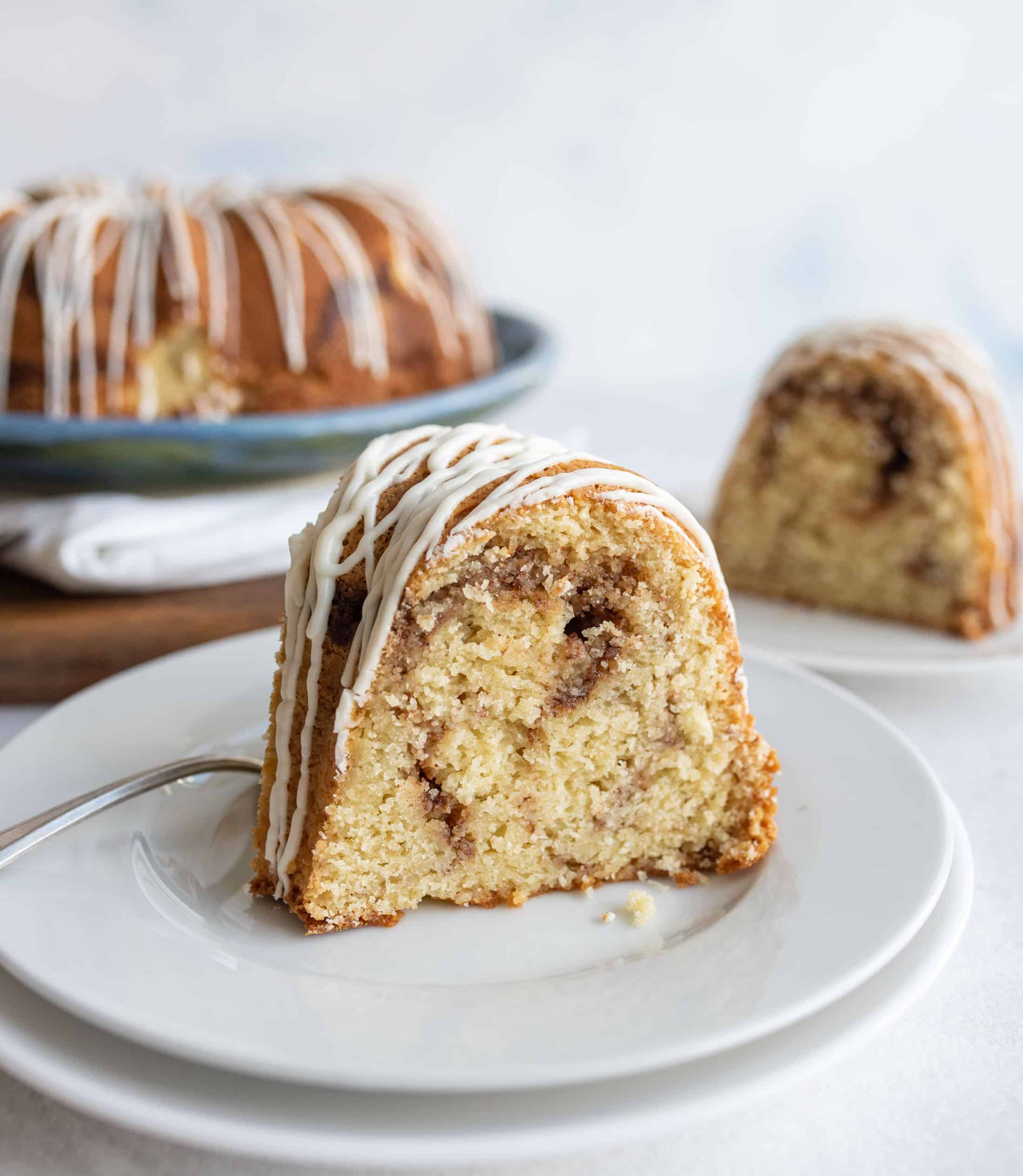 A slice of bundt cake with cinnamon swirl and white icing drizzle sits on a white plate with a fork. More cake is visible in the background on a serving platter and another plate.