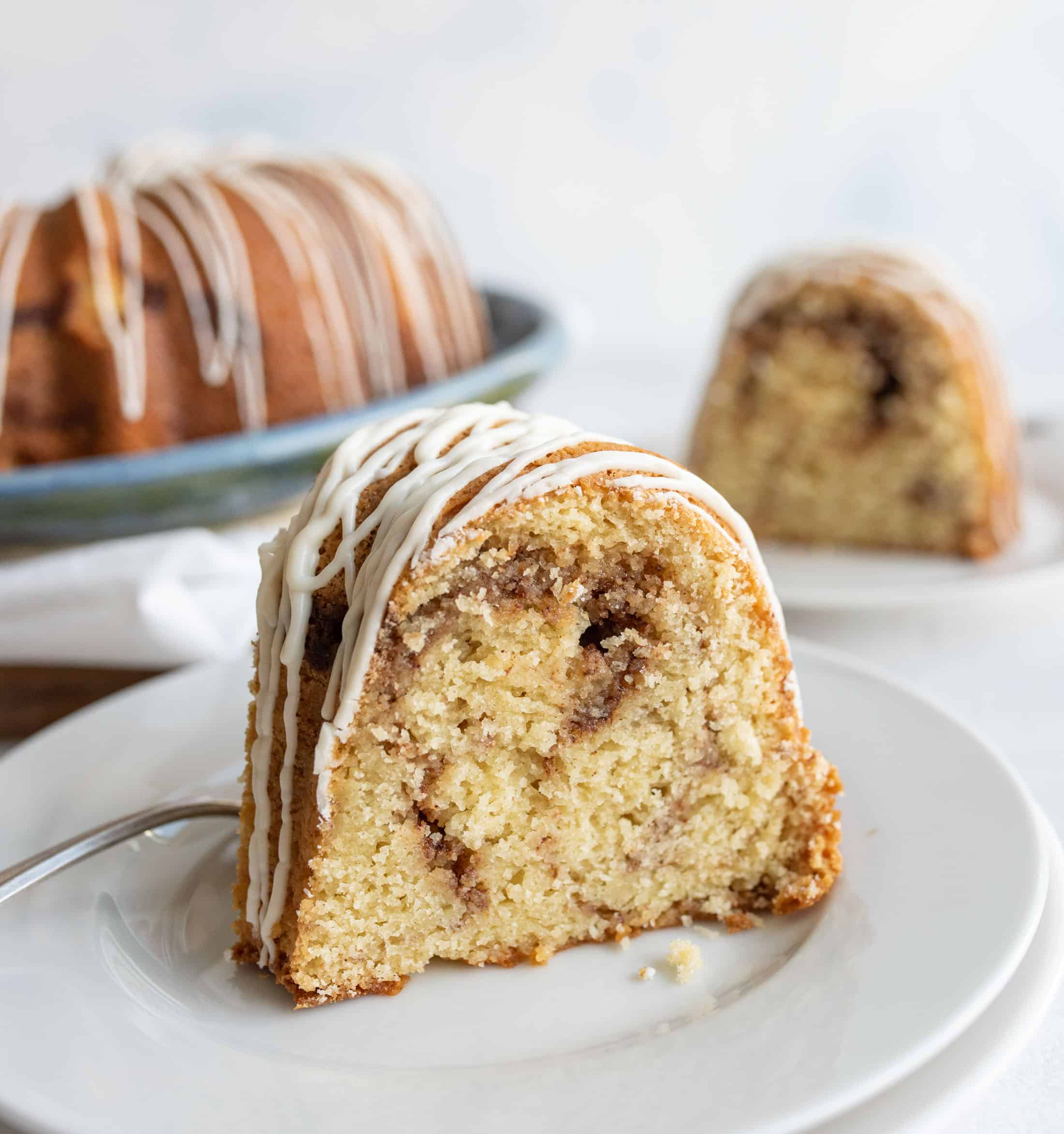 A close-up of a slice of cinnamon swirl bundt cake with white icing drizzle, served on a white plate with a fork. More cake slices are visible in the background.