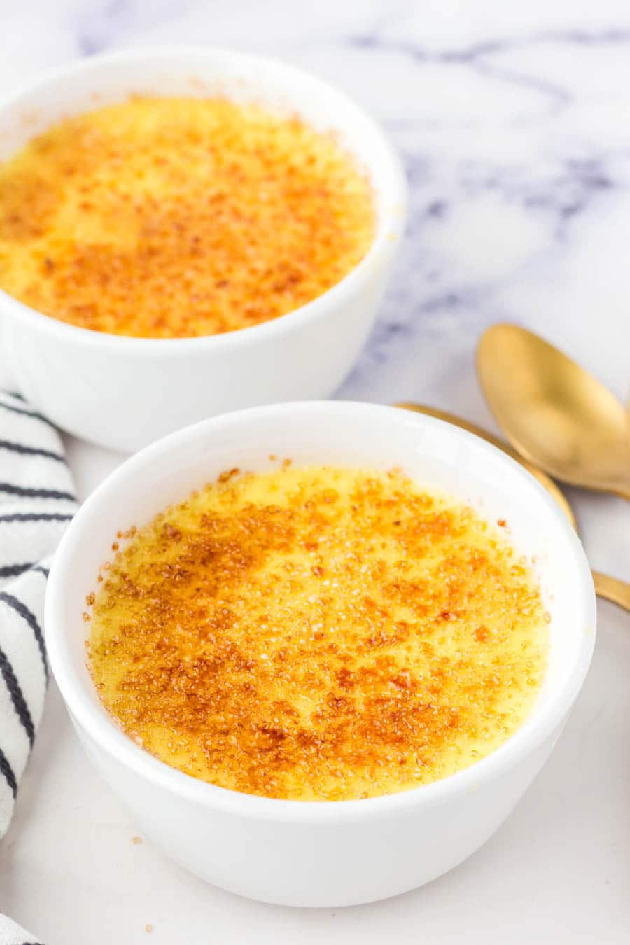 Two white ramekins filled with crème brûlée topped with a golden, caramelized sugar crust, placed on a white surface with two gold spoons and a striped cloth nearby.