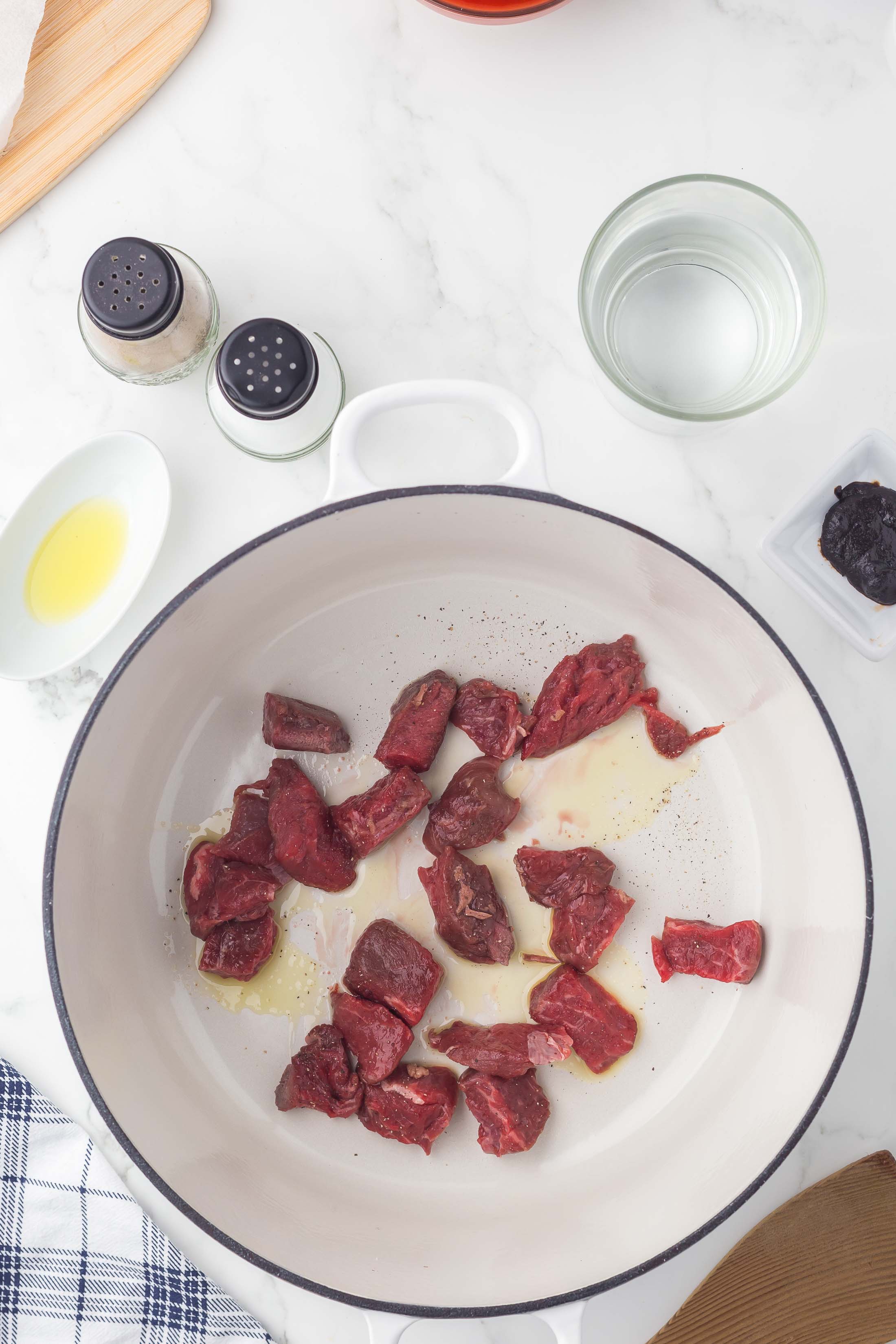 Raw beef chunks in a white pot, surrounded by small bowls of oil, black paste, water, salt and pepper shakers, and a checkered cloth on a marble countertop.