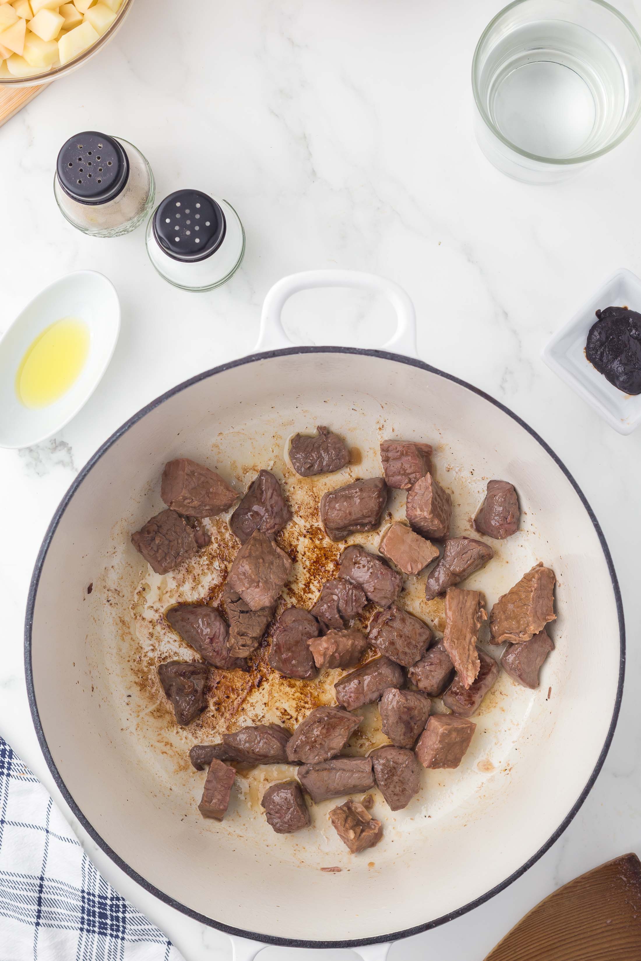 Chunks of browned beef cook in a white pot on a marble counter, surrounded by salt, pepper, a bowl of water, olive oil, diced potatoes, a prune, and a towel.
