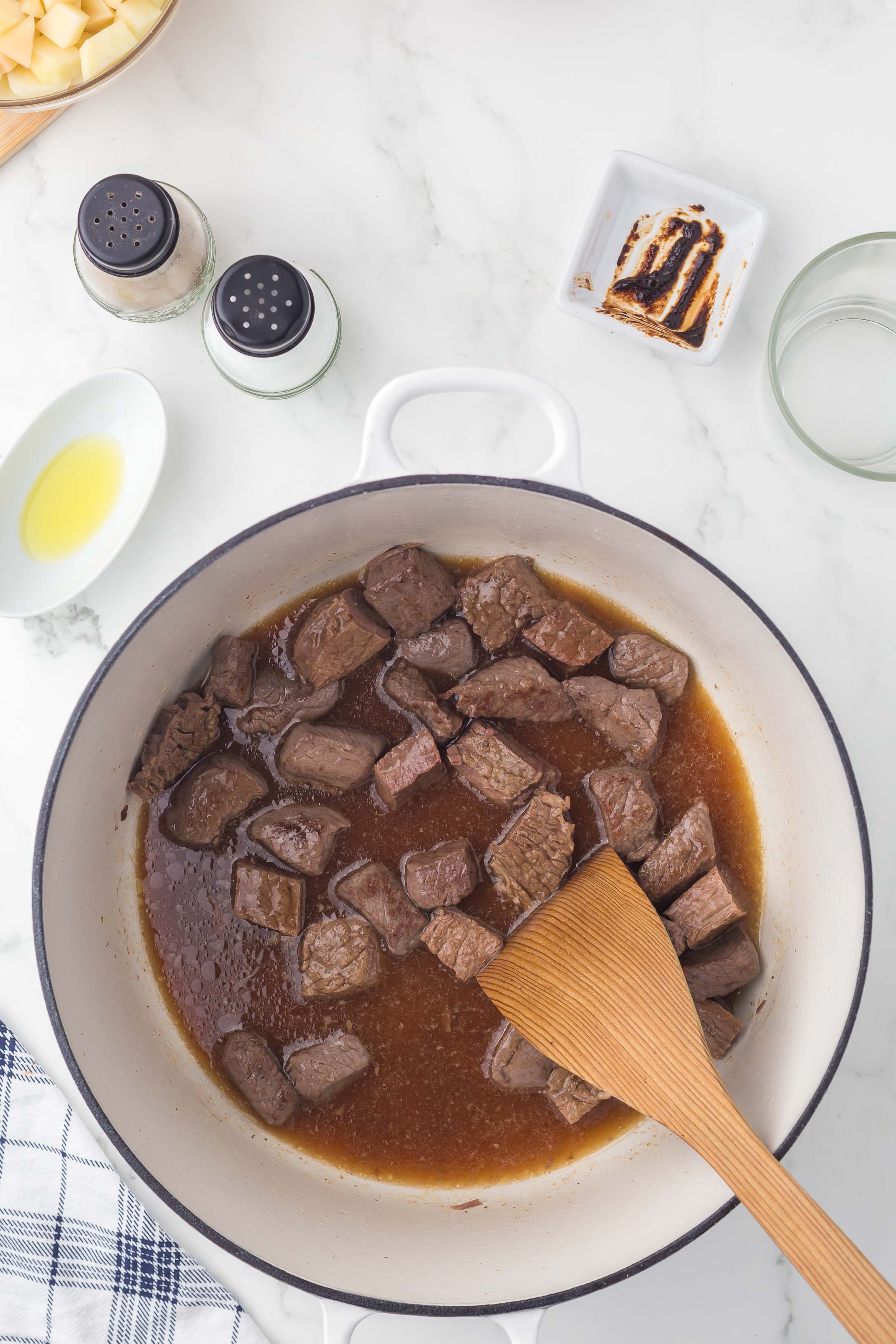 Chunks of beef simmer in brown broth inside a white pot, stirred by a wooden spatula. Surrounding the pot are seasonings, sliced bread with sauce, a bowl of oil, and a checkered towel on a marble countertop.